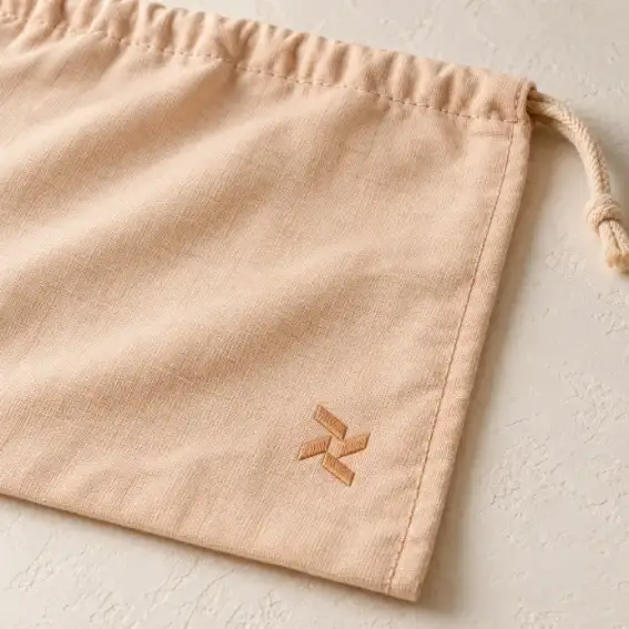 Luxury Dust Bag Logo Customization Detail Pale-apricot cotton dust bag with a small abstract embroidered emblem near the lower-right front panel.